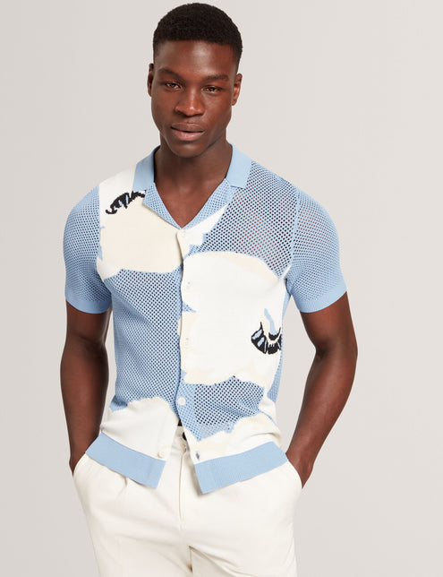 A male model wearing a light blue short sleeve knitted shirt with a large floral graphic and open neck revere collar.