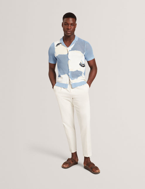 A male model wearing a light blue short sleeve knitted shirt with a large floral graphic and open neck revere collar.