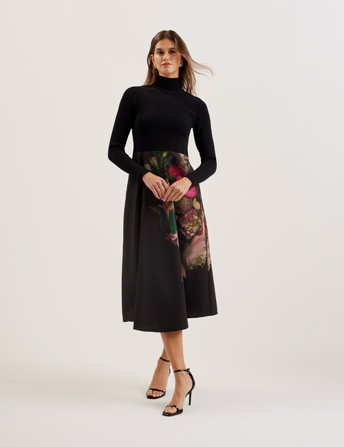 A woman wearing a black midi dress with a high funnel neck, long sleeves, and a wrap style woven skirt with a floral print. She is standing against a white background.